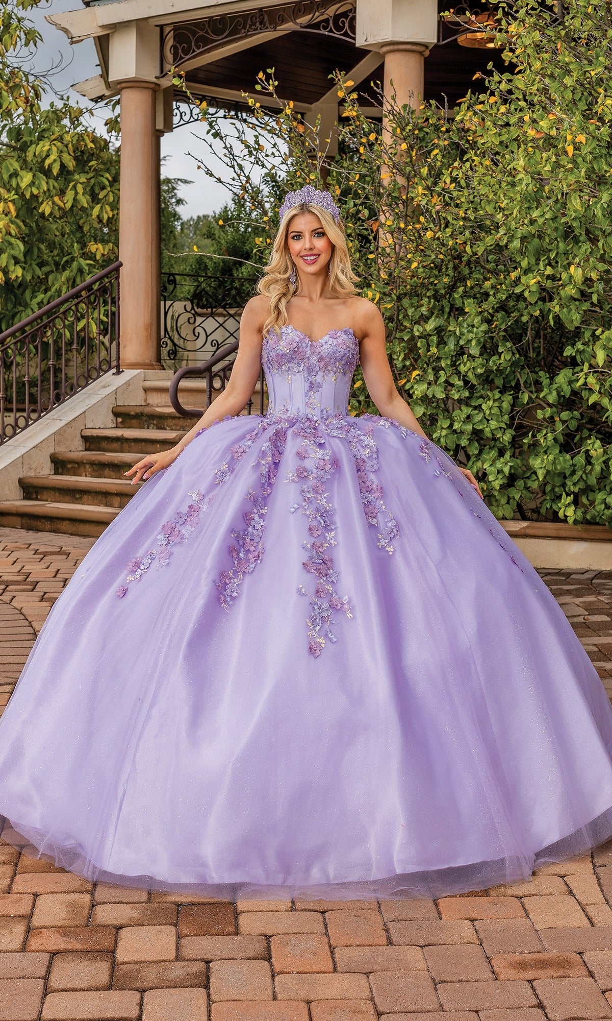 A woman poses outdoors on a brick path in front of greenery and a gazebo, wearing the Quinceanera Dress 1855 By Dancing Queen—a lilac gown with floral embellishments, strapless sweetheart neckline, and tiara that enhance her enchanting look.